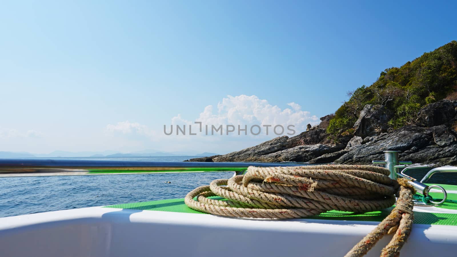 View of the bow of a fast boat with a rope. The metal rim glitters. It offers a landscape on the blue sea, a green island with trees and large stones. In the distance the blue sky with white clouds.