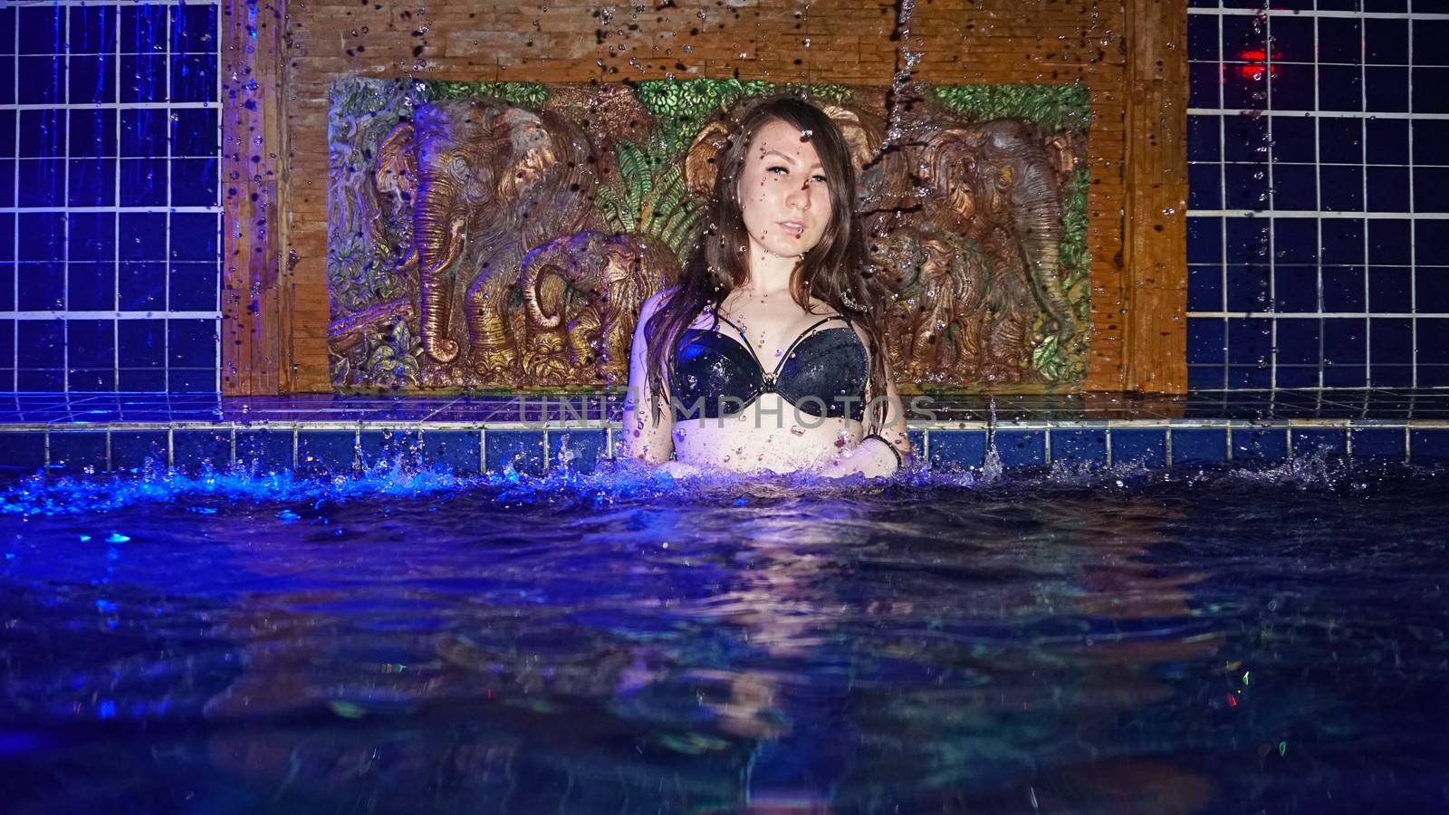 A girl in a black swimsuit and glasses poses on the edge of the pool. Swimming pool on the roof of the building. Photosession. View of the blue sky and clear water. Small waterfall. Thailand, Phuket.