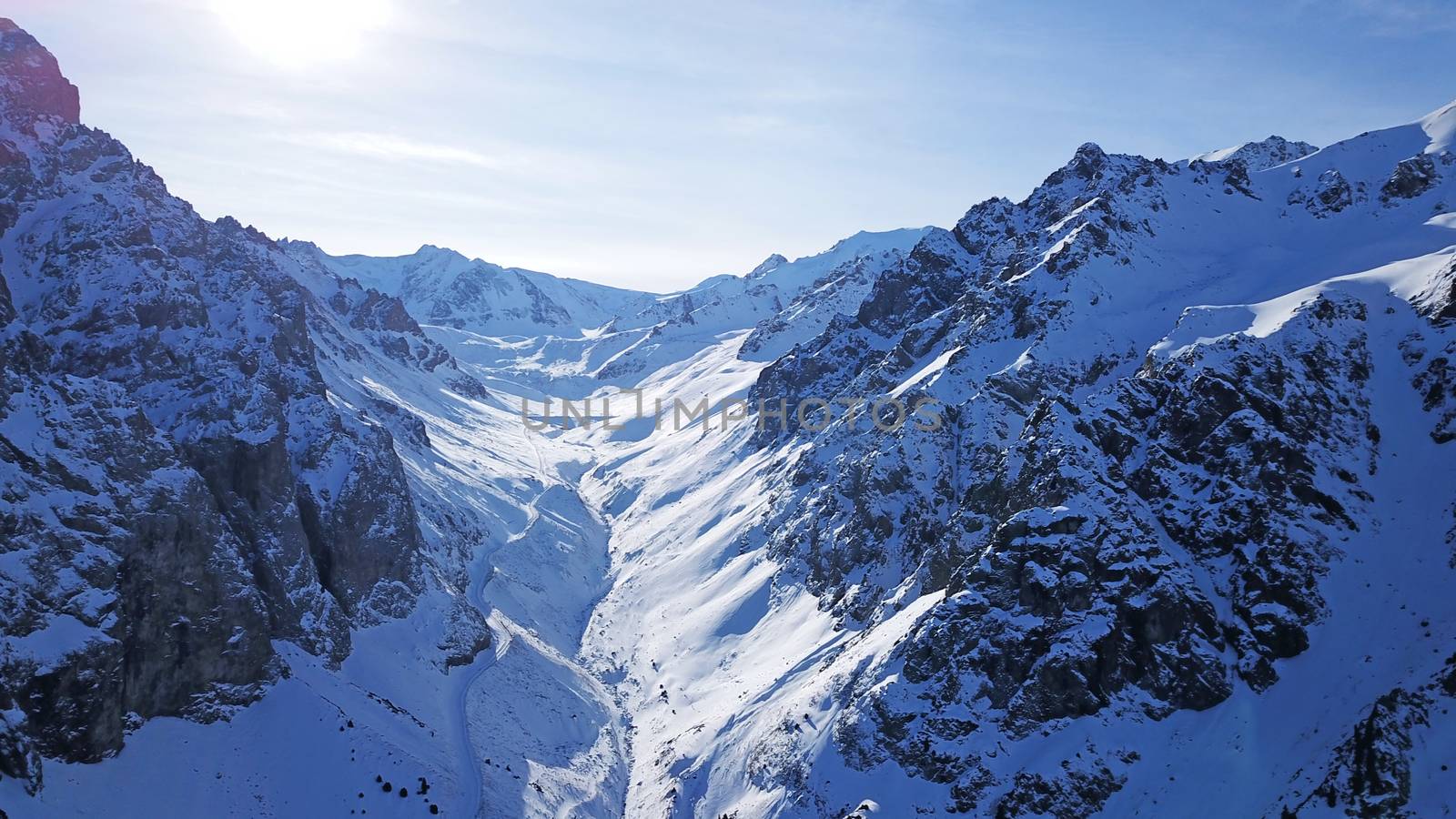 High snowy mountains, spruce trees grow in places. Huge rocks and cliffs, gorges where there may be an avalanche. Top view from a drone. Sunny day, shadows from the mountains fall on the snow. Almaty