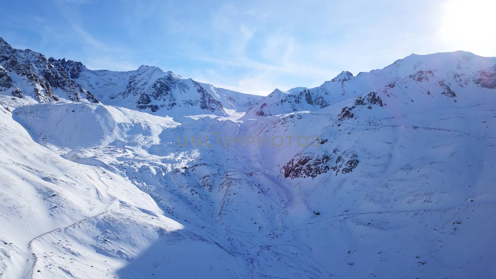 Snow mountain peaks with rocks. View from drone. In places, you can see glaciers, large rocks and rocks. Huge drifts of snow covered mountains. Dangerous place. Blue sky and sun. Shadow of the peaks.