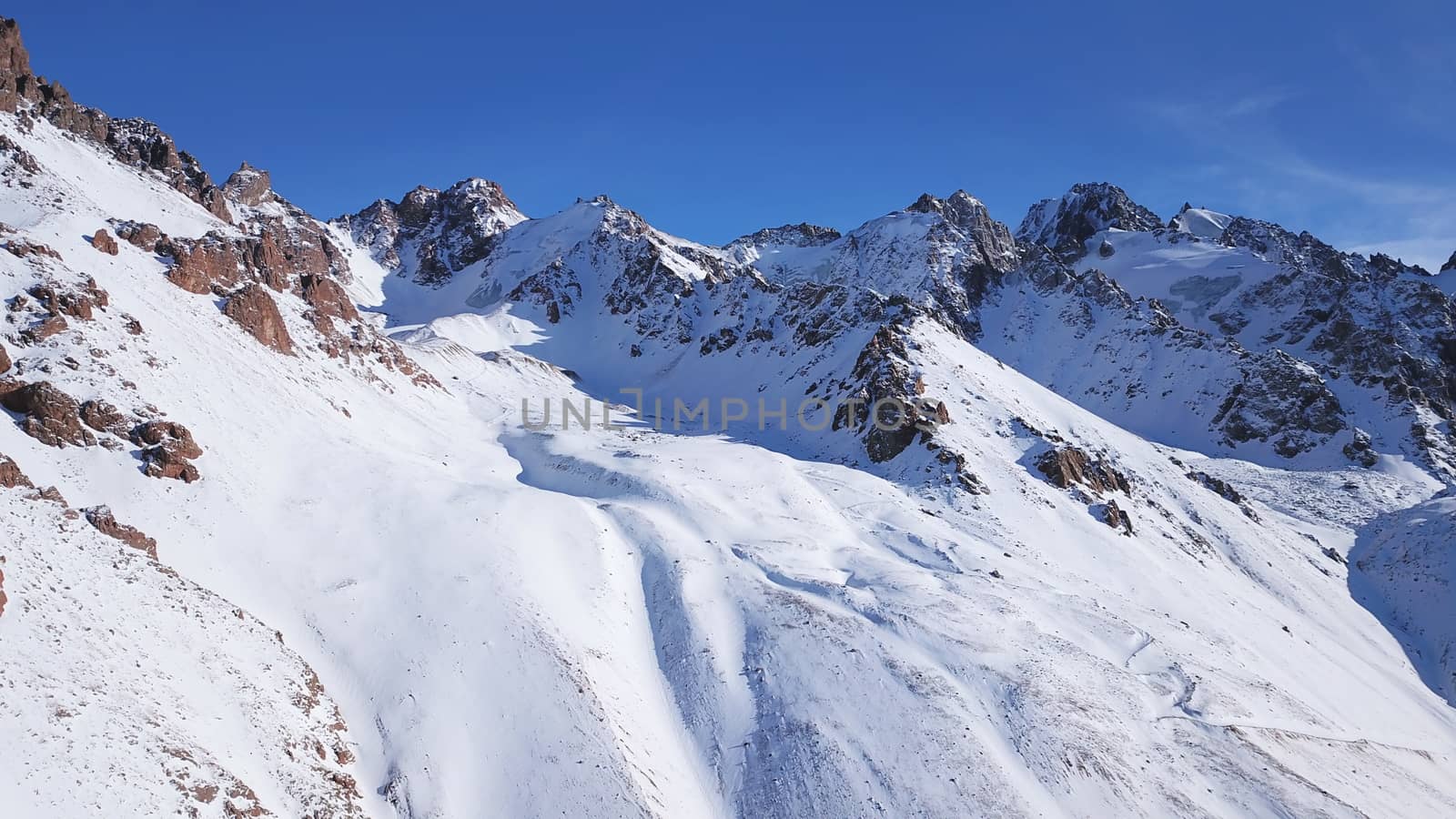 Snow mountain peaks with rocks. View from drone. In places, you can see glaciers, large rocks and rocks. Huge drifts of snow covered mountains. Dangerous place. Blue sky and sun. Shadow of the peaks.