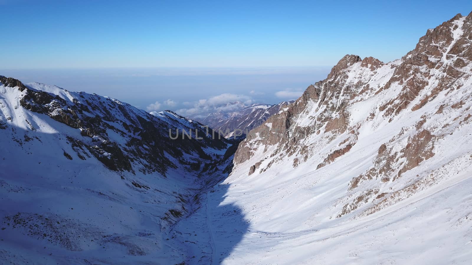 Snow mountain peaks with rocks. View from drone. In places, you can see glaciers, large rocks and rocks. Huge drifts of snow covered mountains. Dangerous place. Blue sky and sun. Shadow of the peaks.