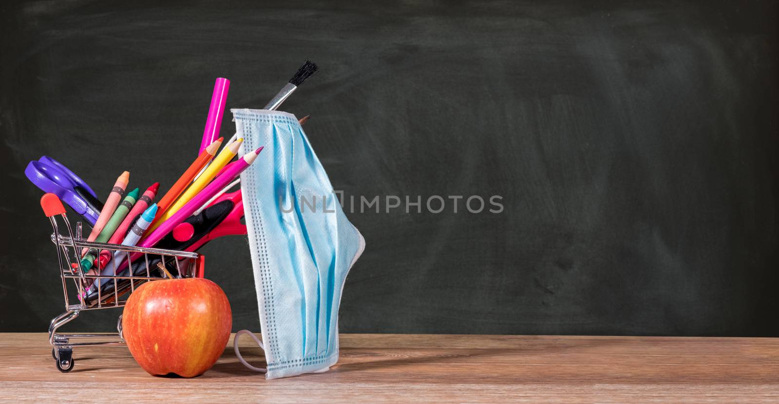 Back to school during coronavirus pandemic with pencils, crayons and face mask in shopping cart with apple against chalkboard