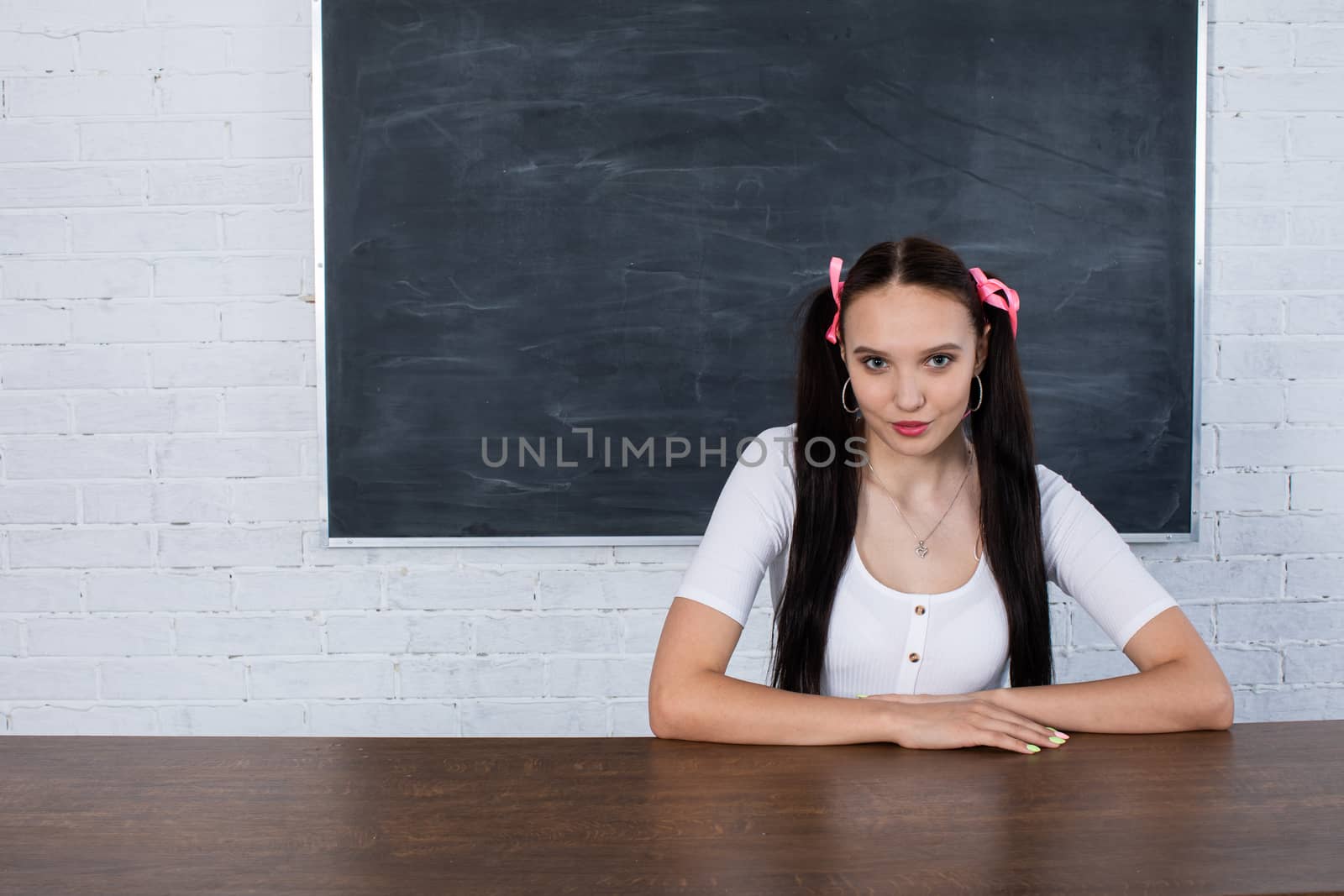 The student is sitting at the school desk waiting for the next lesson to start. A young girl has a hairstyle in ponytails. by fotodrobik