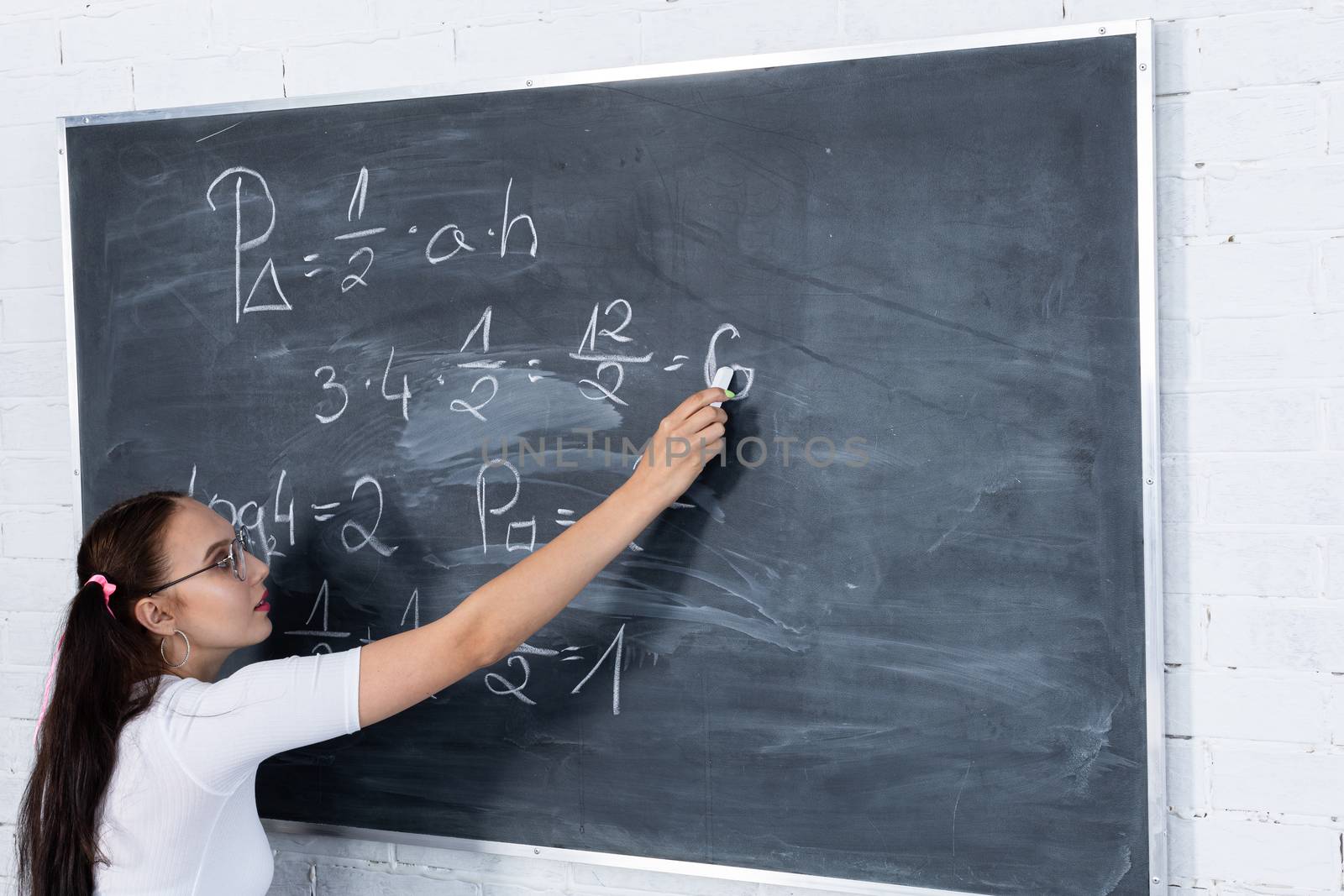 A teenage girl is solving a math problem at school by writing on a school blackboard with white chalk. Her hair is pinned with a pink ribbon in two ponytails. by fotodrobik