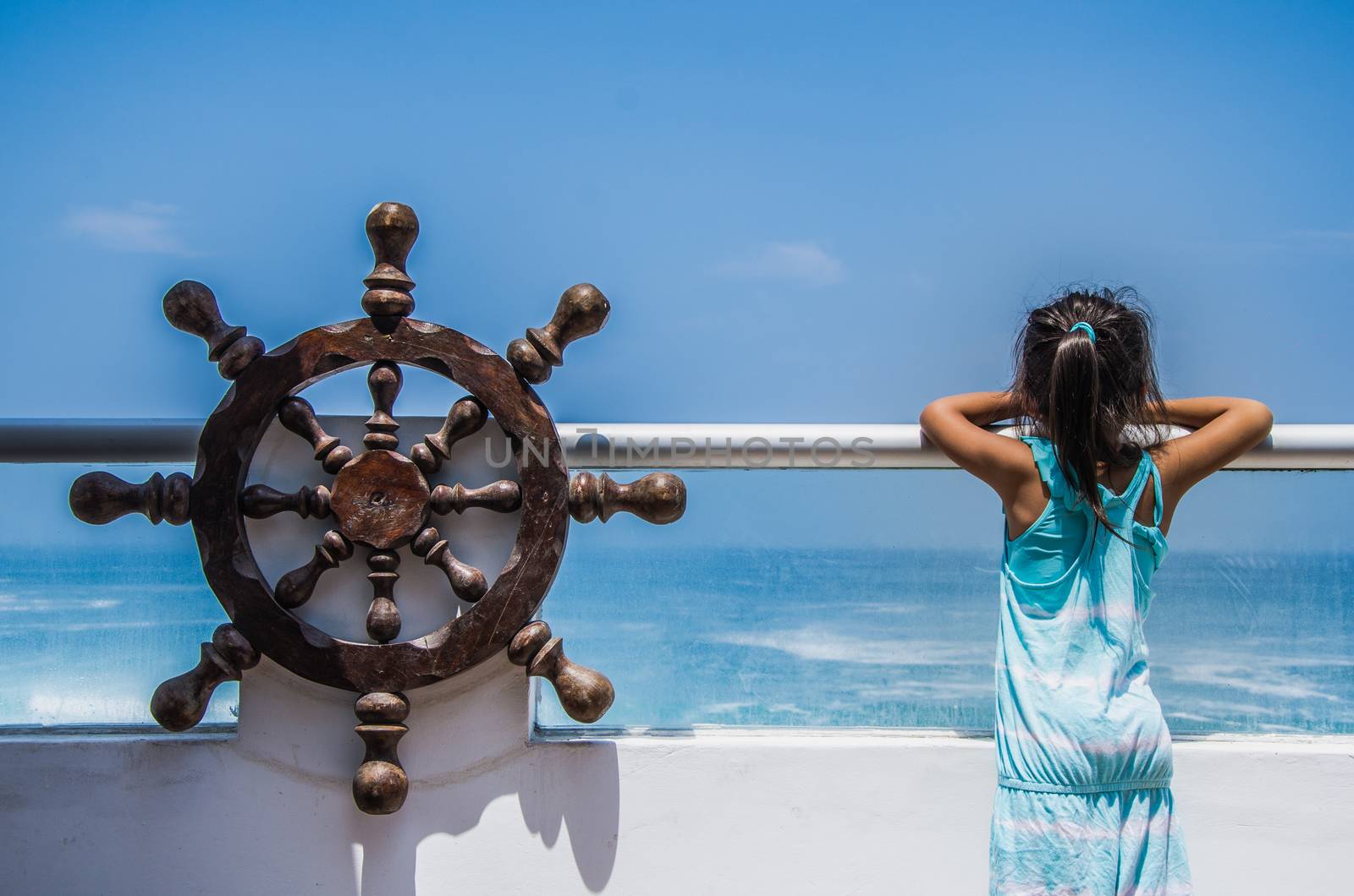 Girl observing the sea from a balcony of a house on the beach with a pirate ship helm at her side, enjoying life and summer vacation, rear view