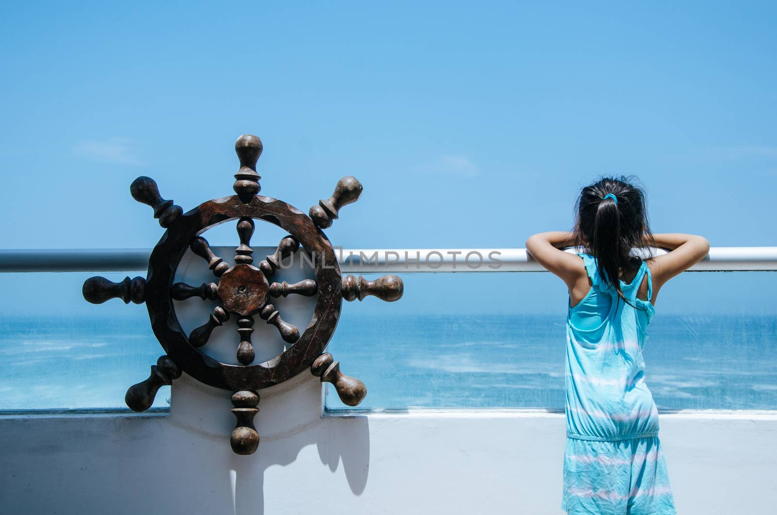 Girl observing the sea from a balcony of a house on the beach with a pirate ship helm at her side, enjoying life and summer vacation, rear view