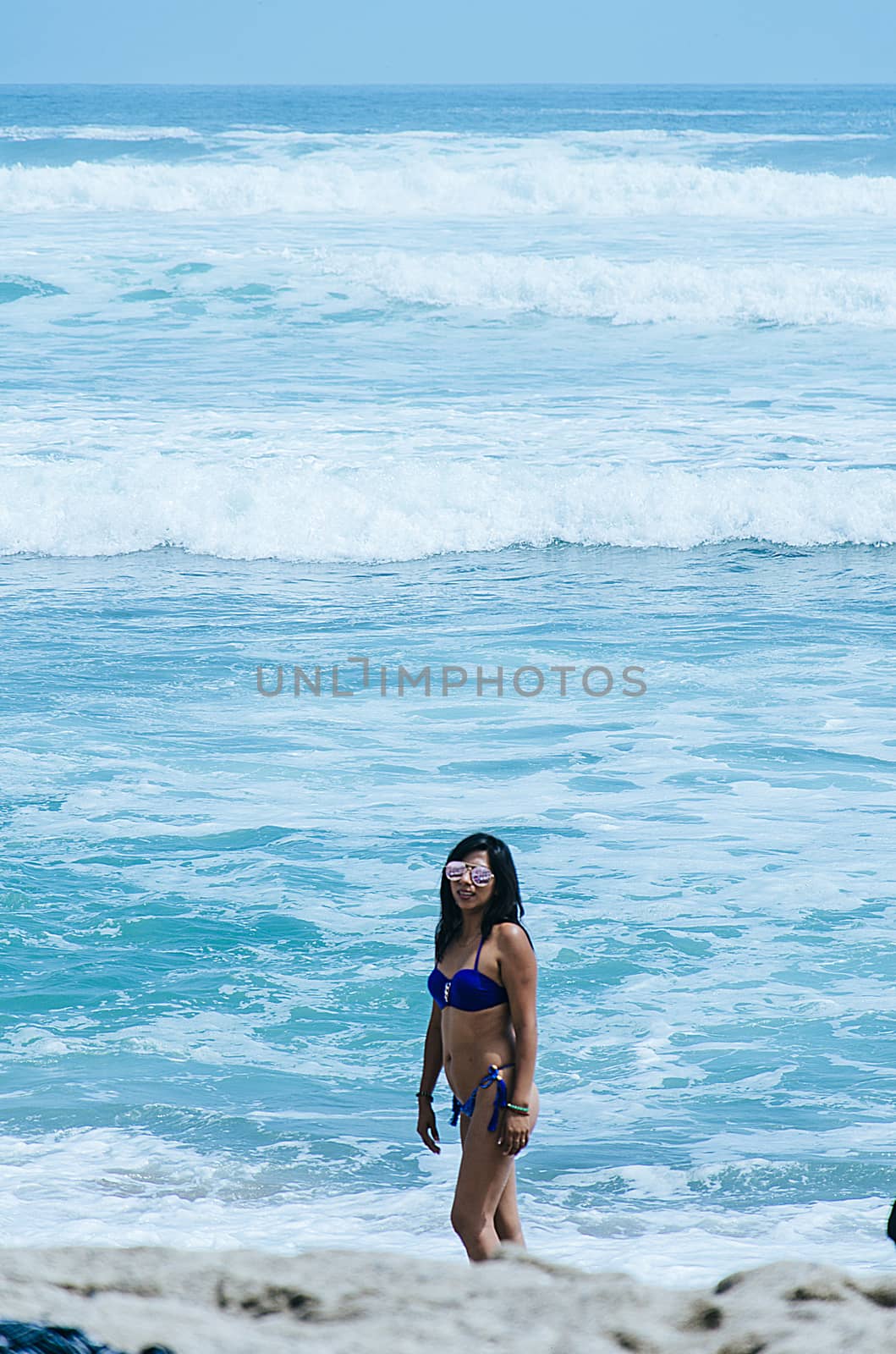 Young fashion seductive girl in colorful stylish swimwear posing on the beach background. Surprised face. Trendy sunglasses. Perfect tamed slim body, wavy hairstyle.