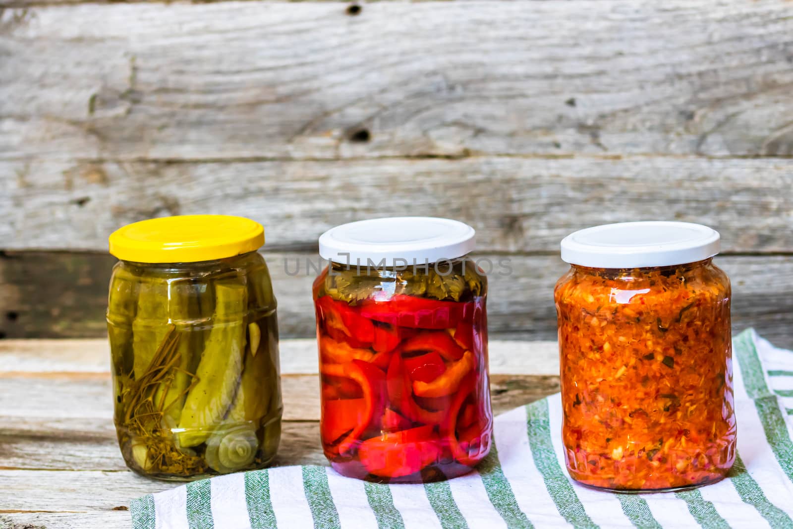 Glass jars with pickled red bell peppers and pickled cucumbers (pickles) isolated. Jars with variety of pickled vegetables. Preserved food concept in a rustic composition.