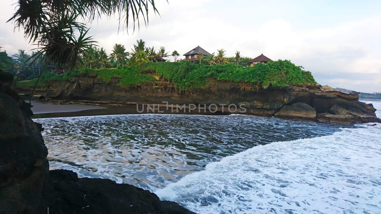 On the edge of the cliff are houses, the rocks are covered with palm trees and greenery. The earth is washed by large waves of the ocean. A Paradise on the island of Bali.