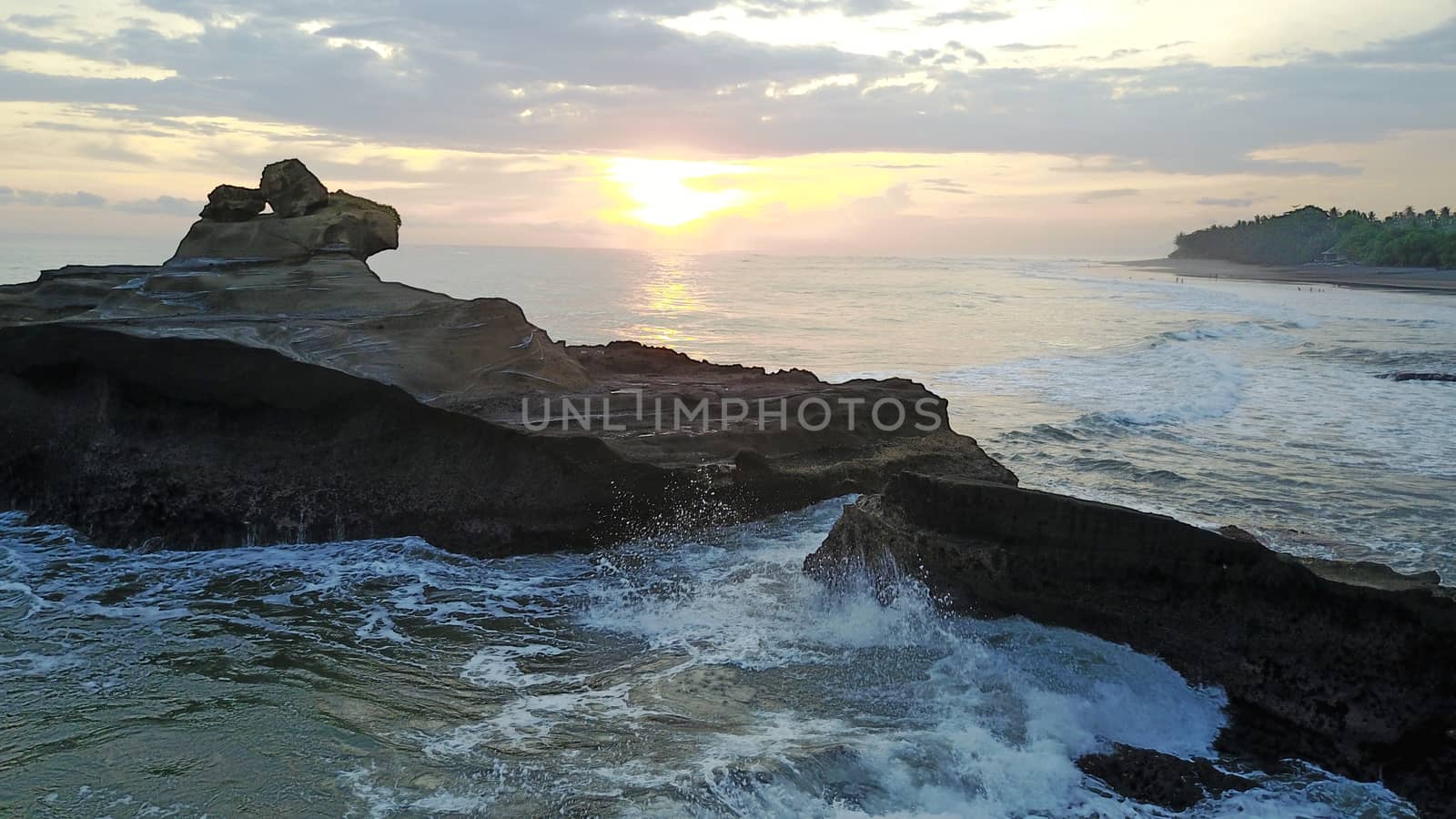 Sunset over the ocean. View of large waves breaking on the rocks. Clouds hang over the horizon. A ray from the sun is reflected on the water in orange color. The coast of Bali is visible.