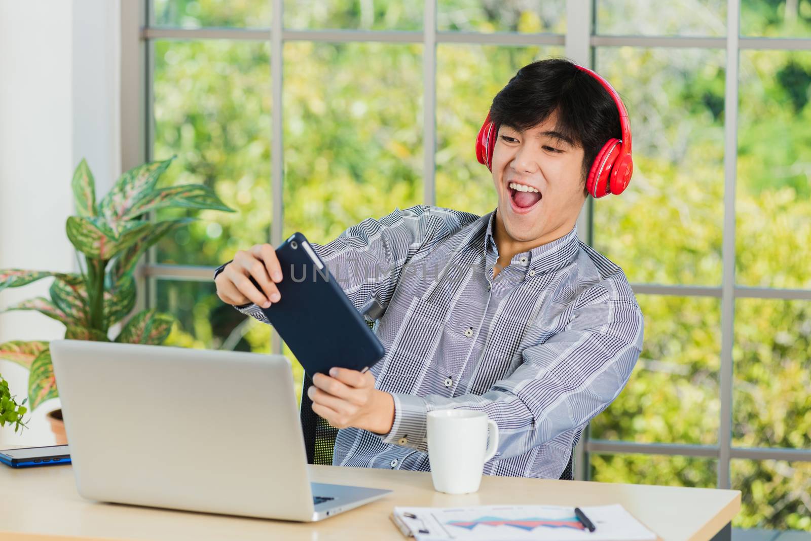 Asian young business man with red headphones he sitting on desk workplace using digital tablet play racing online video games, confident handsome man lifestyle he smiles and funny