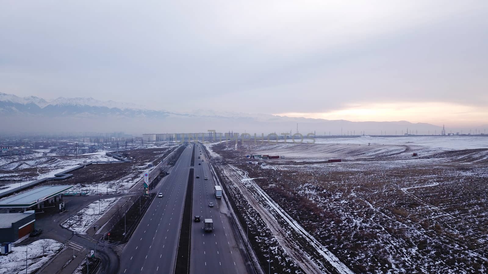 A large truck is traveling on a multi-lane highway. Around the steppe, snow and emptiness. Cloudy weather, winter. The trucker continues on his way. Cars overtake. Transport company. Kazakhstan.