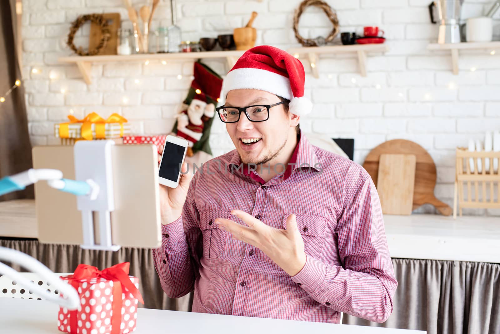 Christmas online greetings. Happy young man in santa hat greeting his friends in video chat or call on tablet sitting in his kitchen