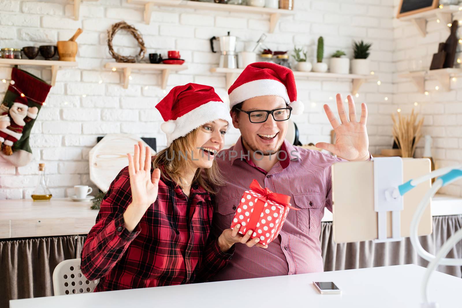 Christmas online greetings. Happy young couple in santa hats greeting their friends in a video call on tablet sitting at their kitchen