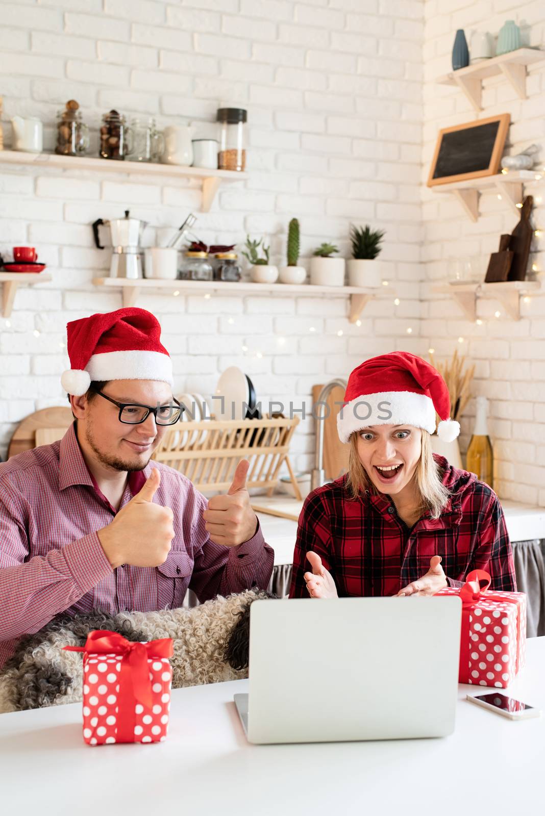 Christmas online greetings. Excited young couple in santa hats greeting their friends in a video call on laptop sitting at the kitchen