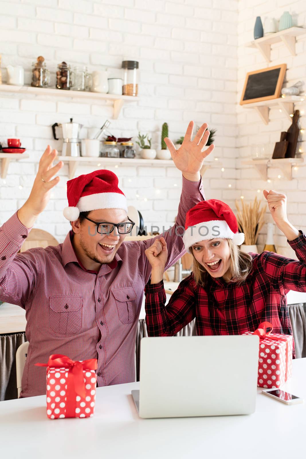 Christmas online greetings. Excited young couple in santa hats greeting their friends in a video call on laptop sitting at the kitchen