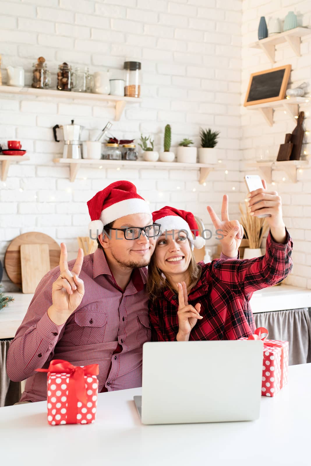 Happy young couple in santa hats greeting their friends in a video call on the laptop and mobile phone by Desperada