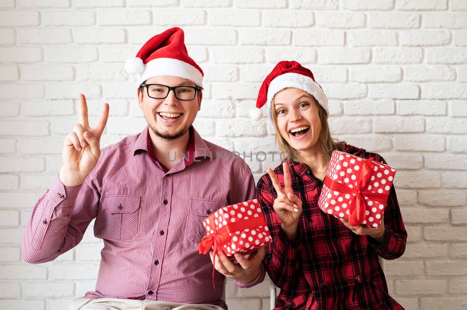 Happy christmas couple in santa hats holding gift boxes on white brick wall background