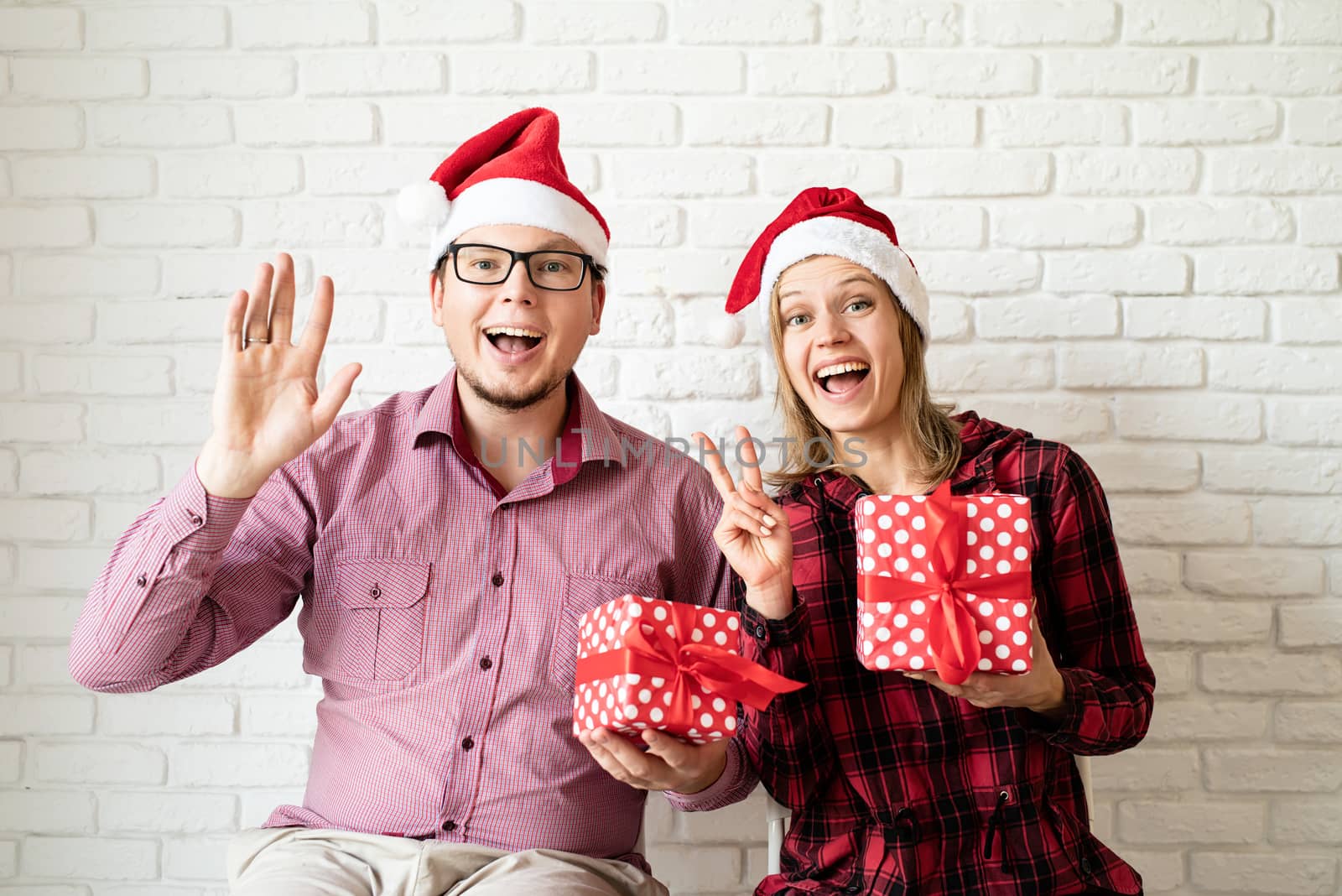 Happy christmas couple in santa hats holding gift boxes on white brick wall background