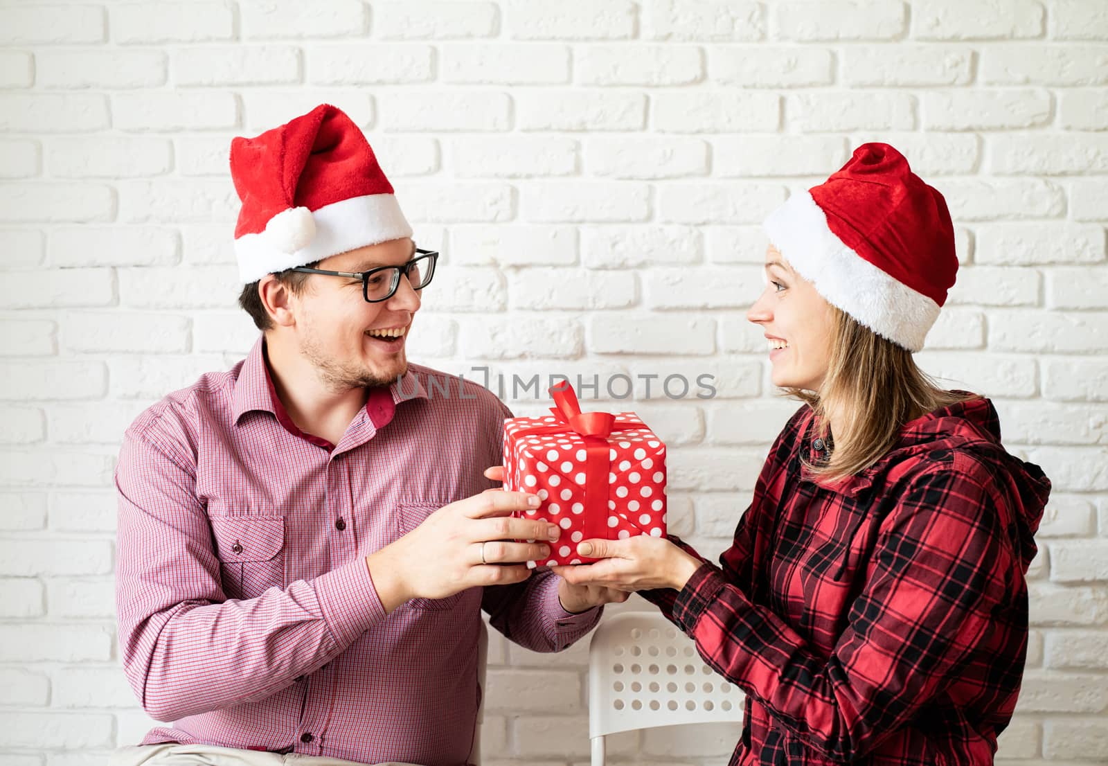 Happy christmas couple in santa hats holding gift boxes on white brick wall background