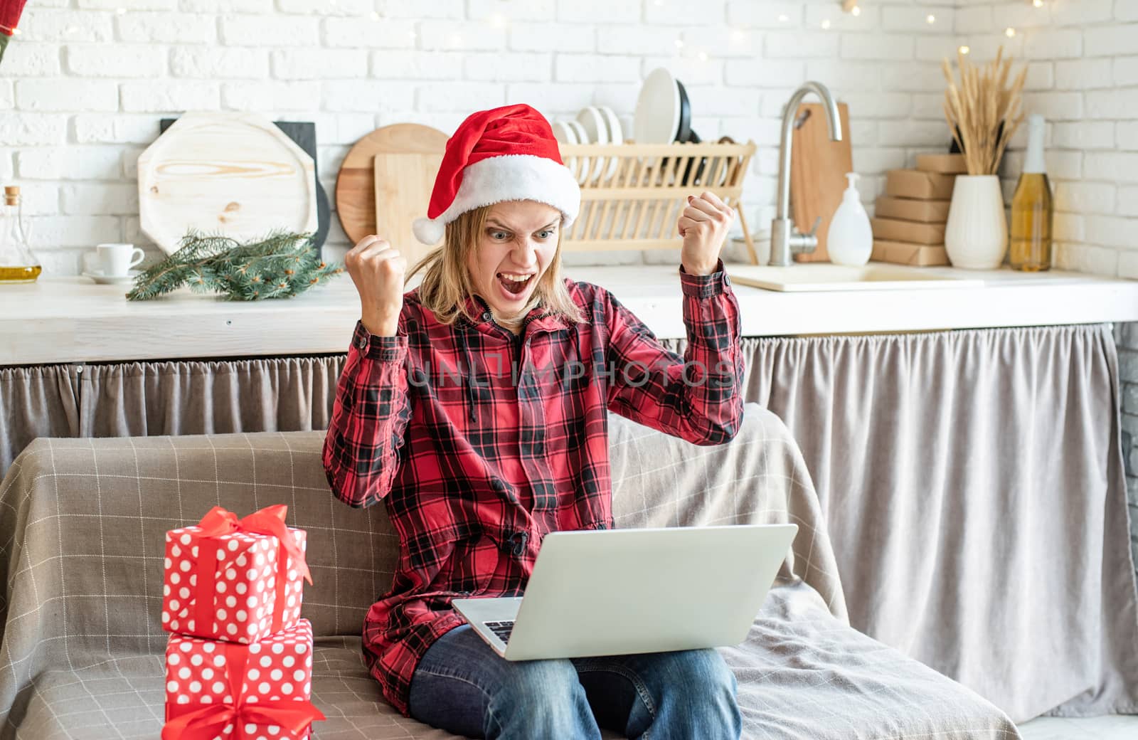Chhristmas online greetings. Excited young woman in santa hat screaming raising her hands working at the laptop