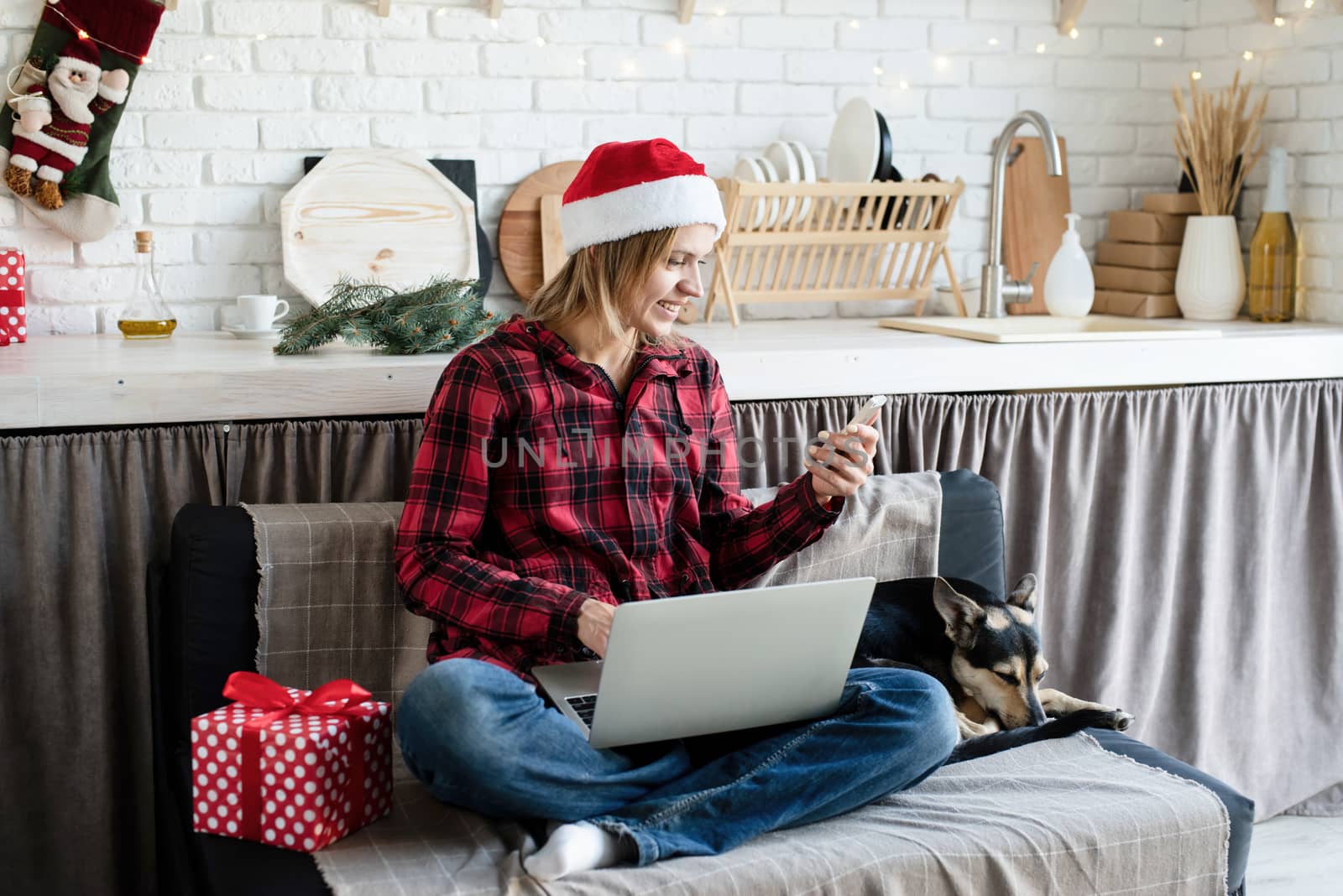 young blond woman in santa hat working on laptop looking at phone by Desperada