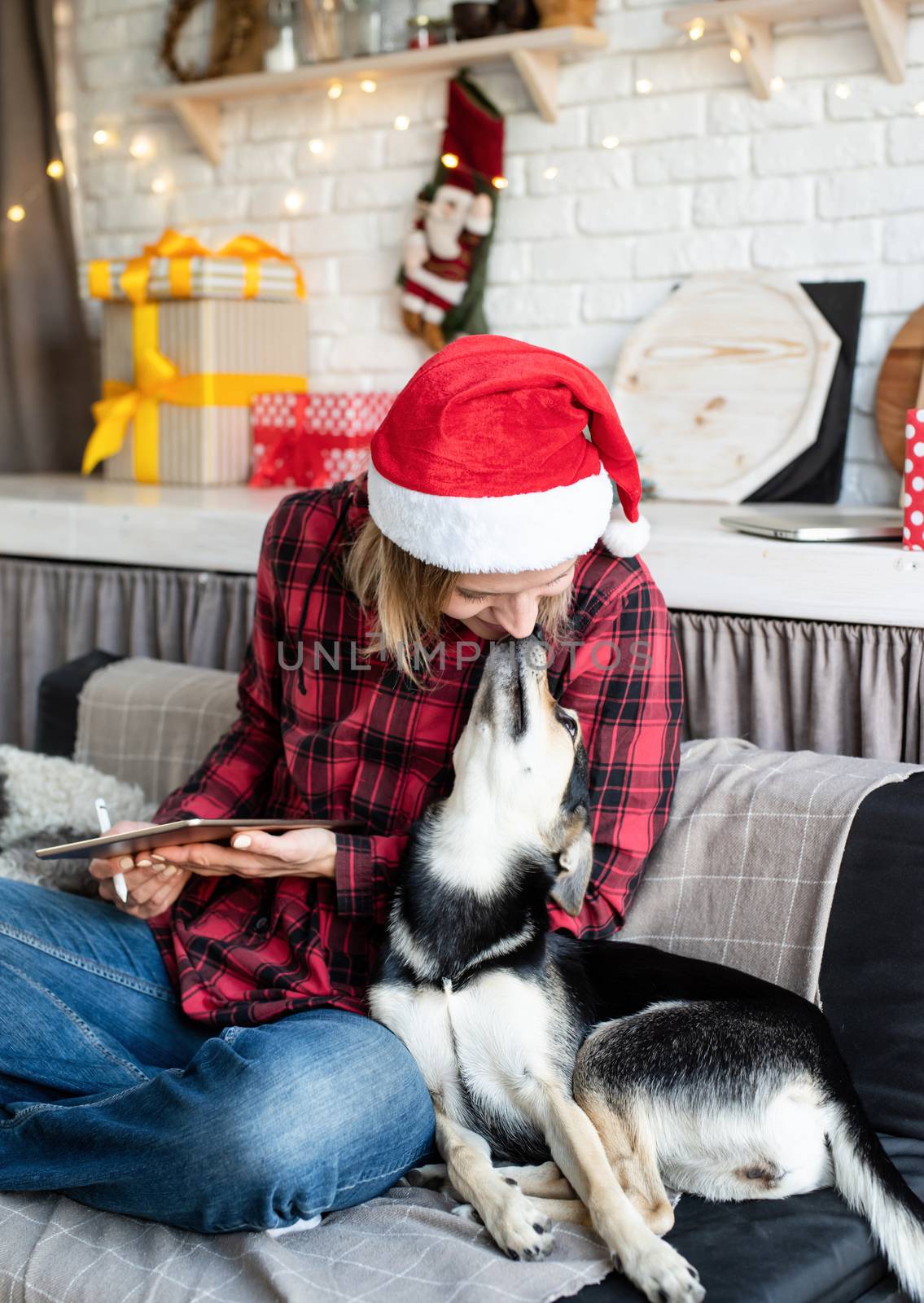 young blond woman in santa hat working on tablet sitting on the couch with her dog by Desperada