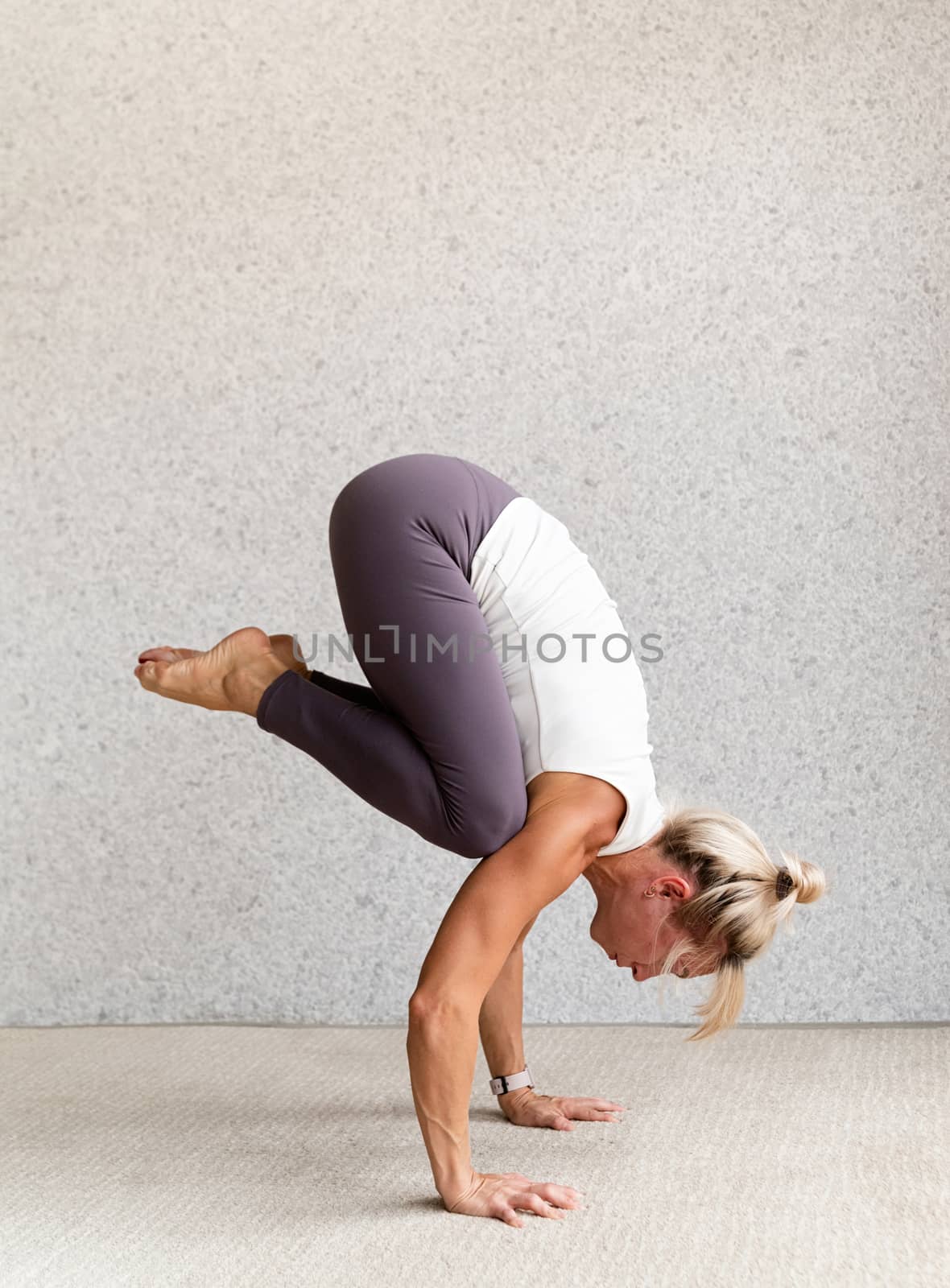 Young attractive woman practicing yoga, wearing sportswear, white shirt and purple pants, indoor full length, gray background by Desperada