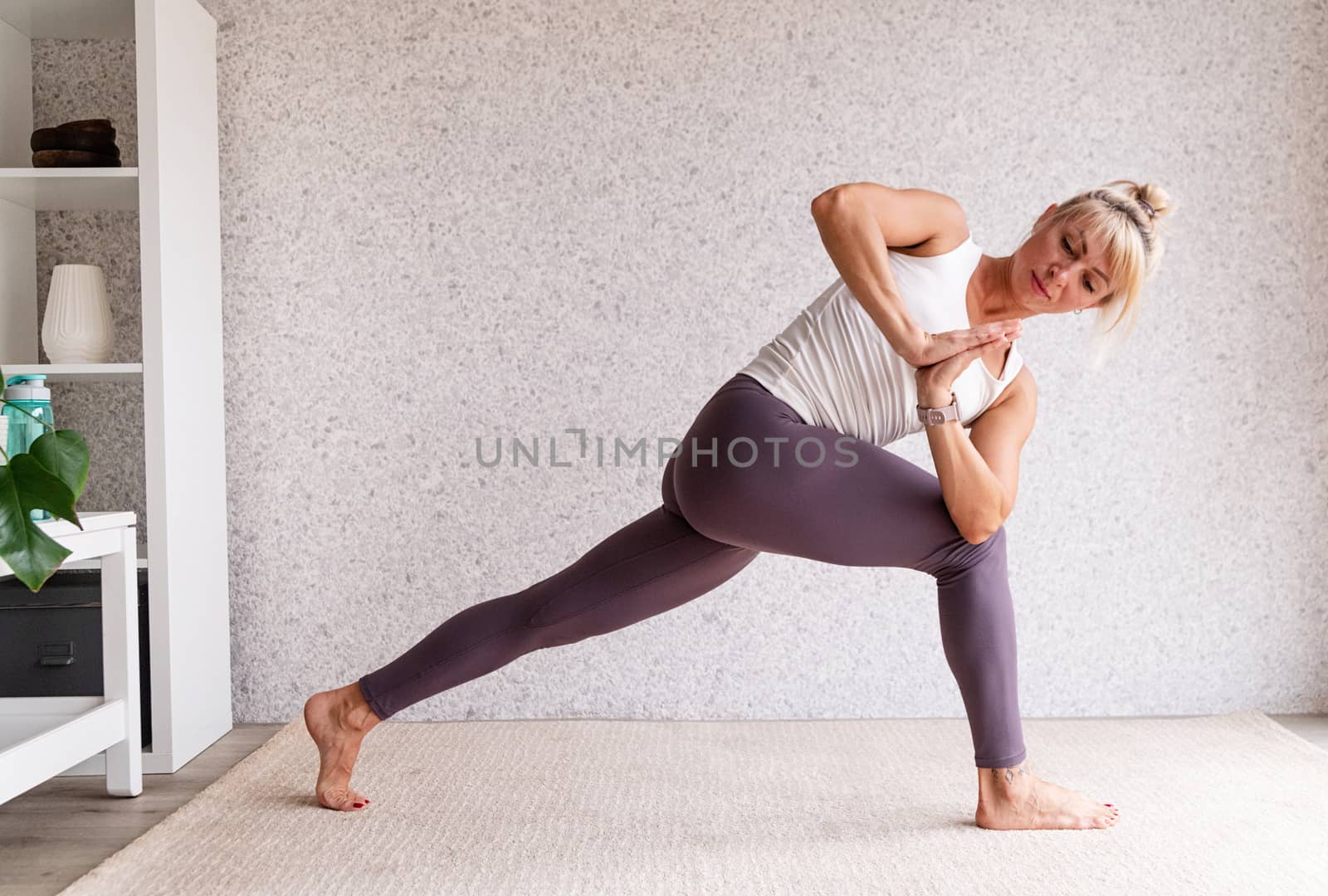 Young attractive woman practicing yoga, wearing sportswear, white shirt and purple pants, indoor full length, gray background by Desperada