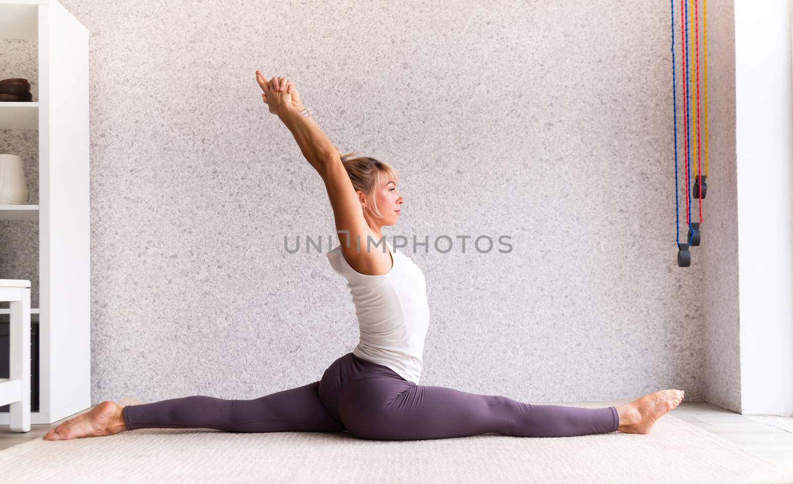 Young attractive woman practicing yoga, wearing sportswear, white shirt and purple pants, indoor full length, gray background by Desperada