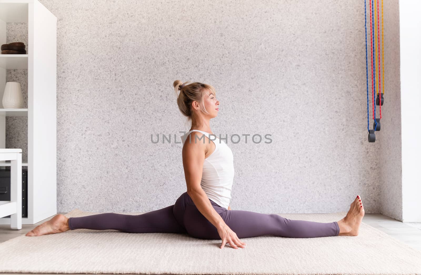 Young attractive woman practicing yoga, wearing sportswear, white shirt and purple pants, indoor full length, gray background by Desperada