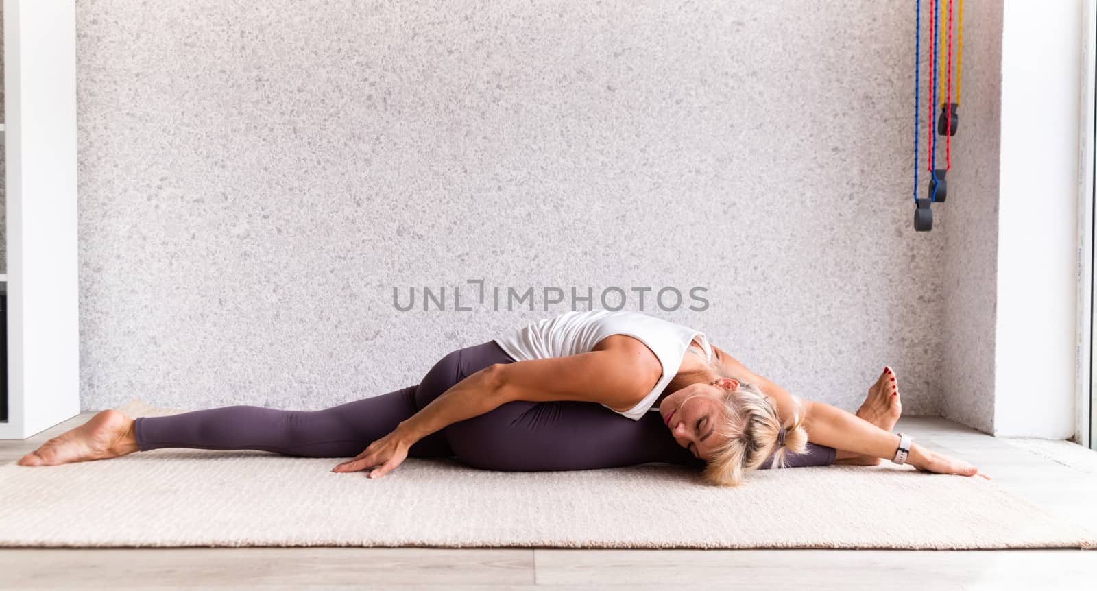 Young attractive woman practicing yoga, wearing sportswear, white shirt and purple pants, indoor full length, gray background by Desperada