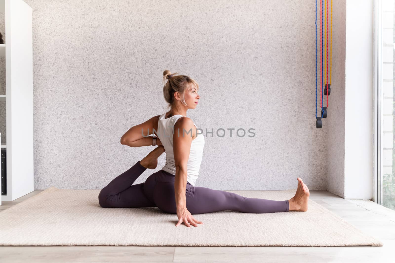 Young attractive woman practicing yoga, wearing sportswear, white shirt and purple pants, indoor full length, gray background by Desperada