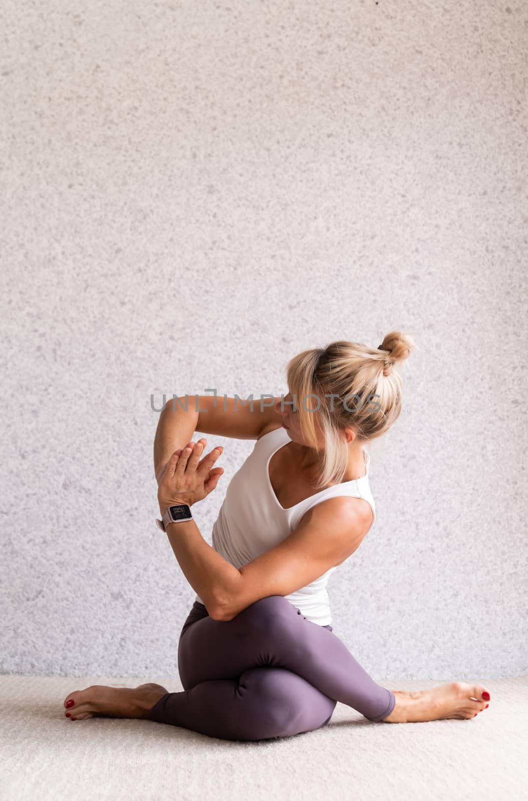 Young attractive woman practicing yoga, wearing sportswear, white shirt and purple pants, indoor full length, gray background by Desperada