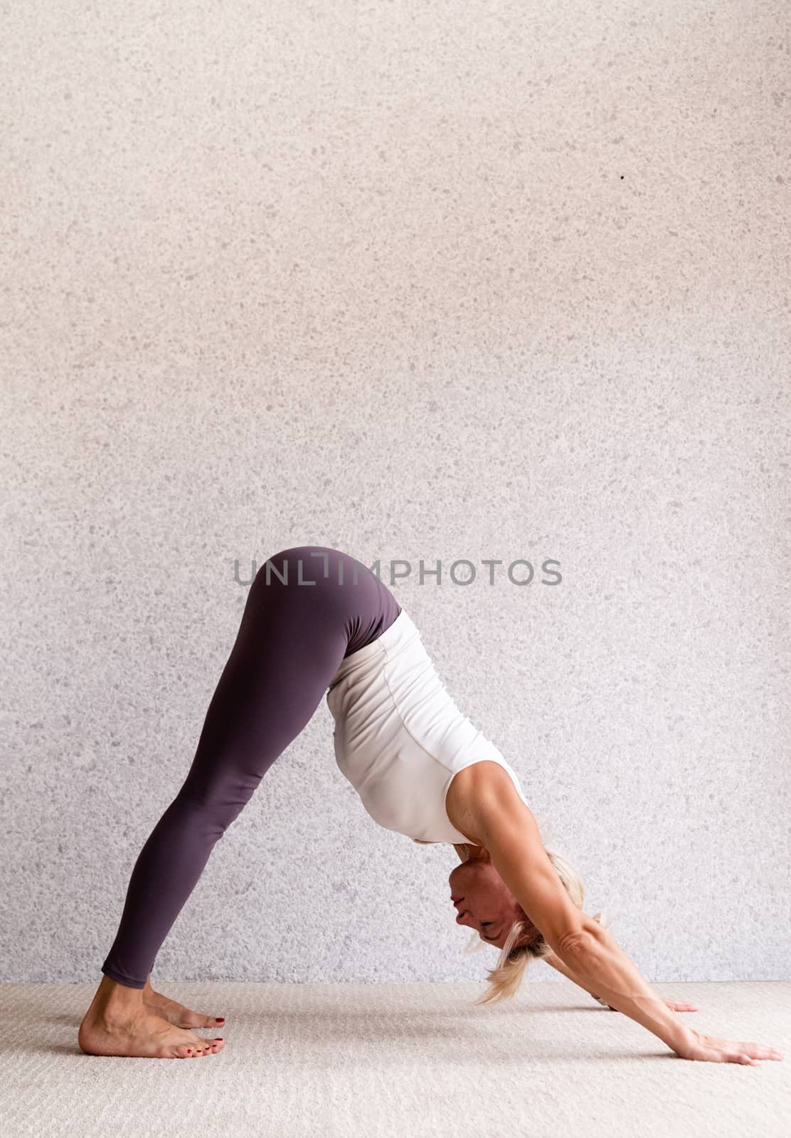 Young attractive woman practicing yoga, wearing sportswear, white shirt and purple pants, indoor full length, gray background by Desperada