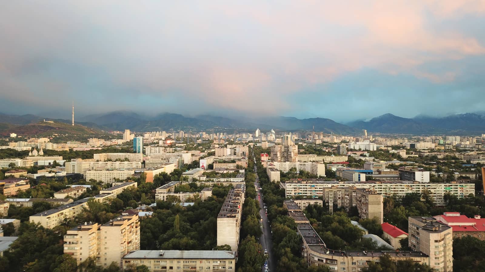 Bright color sunset over the city of Almaty. Huge clouds over the mountains and the city shimmer from bright blue to yellow and dark blue. Tall houses and green trees, cars driving on the roads.