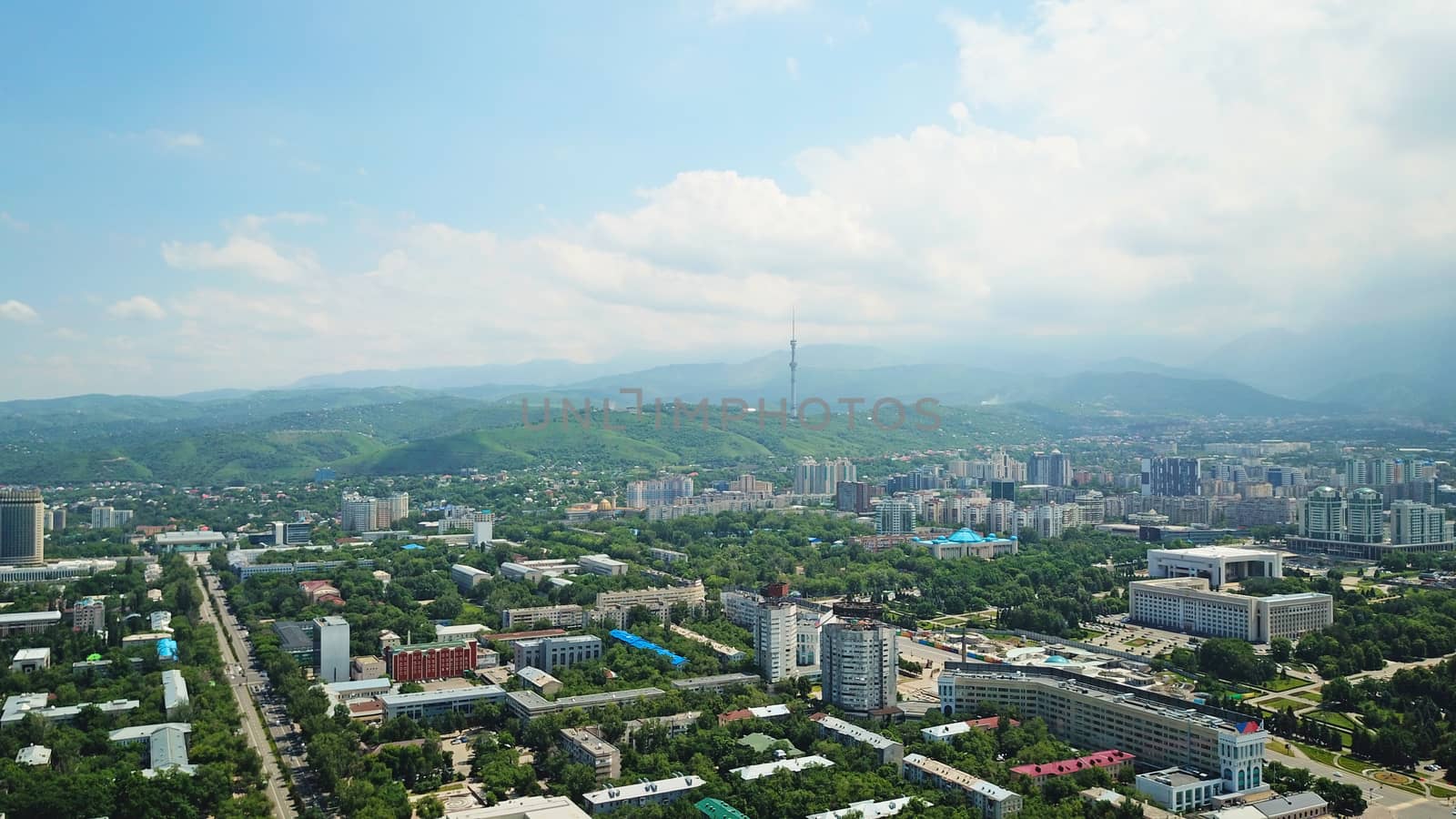 View of Almaty city with white clouds and blue sky. The green city is completely covered with trees, clean roads, transport, houses. On the hill stands the Kok Tobe TV tower. Mountains in the distance