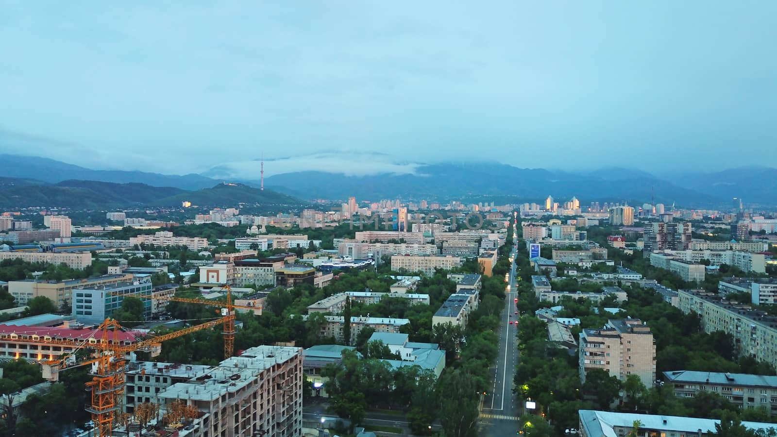 Clouds over the mountains and the city of Almaty at sunset. A lot of green trees, cars driving on the road, high mountains can be seen in the distance. On the hill stands the Kok Tobe TV tower.