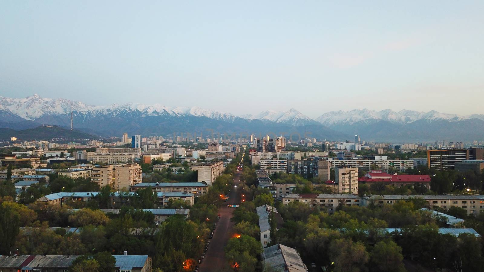 View of the snowy mountains of the TRANS-ili Alatau and the city of Almaty. Clear blue sky, lights are on in the city, cars are driving on the road. Courtyards and streets of the city are illuminated