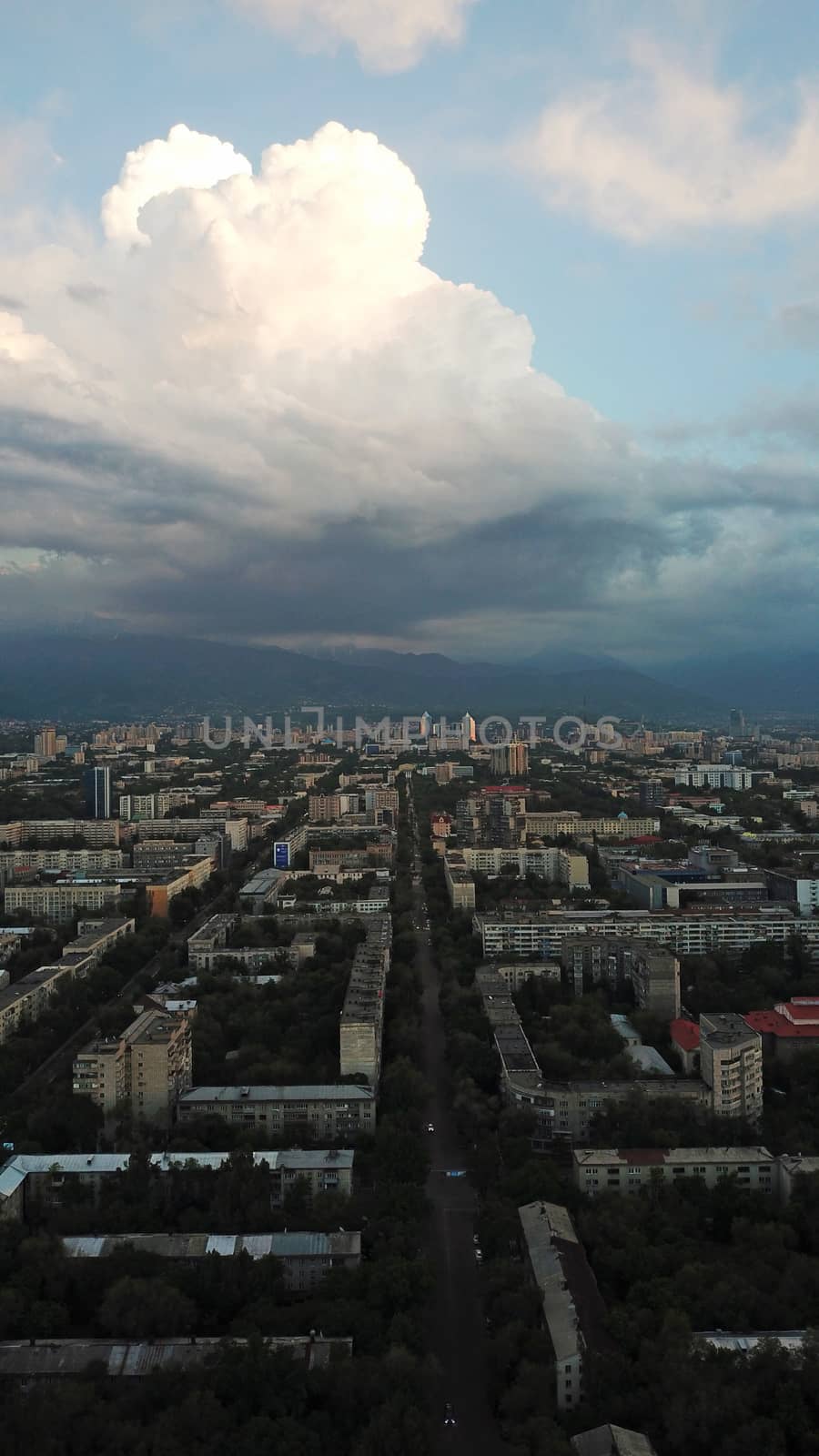 Huge clouds over the city of Almaty. Clouds hang near the mountains. Sunset. You can see different buildings, mosques, churches, and parks. Everything is illuminated by the red rays of the sun.