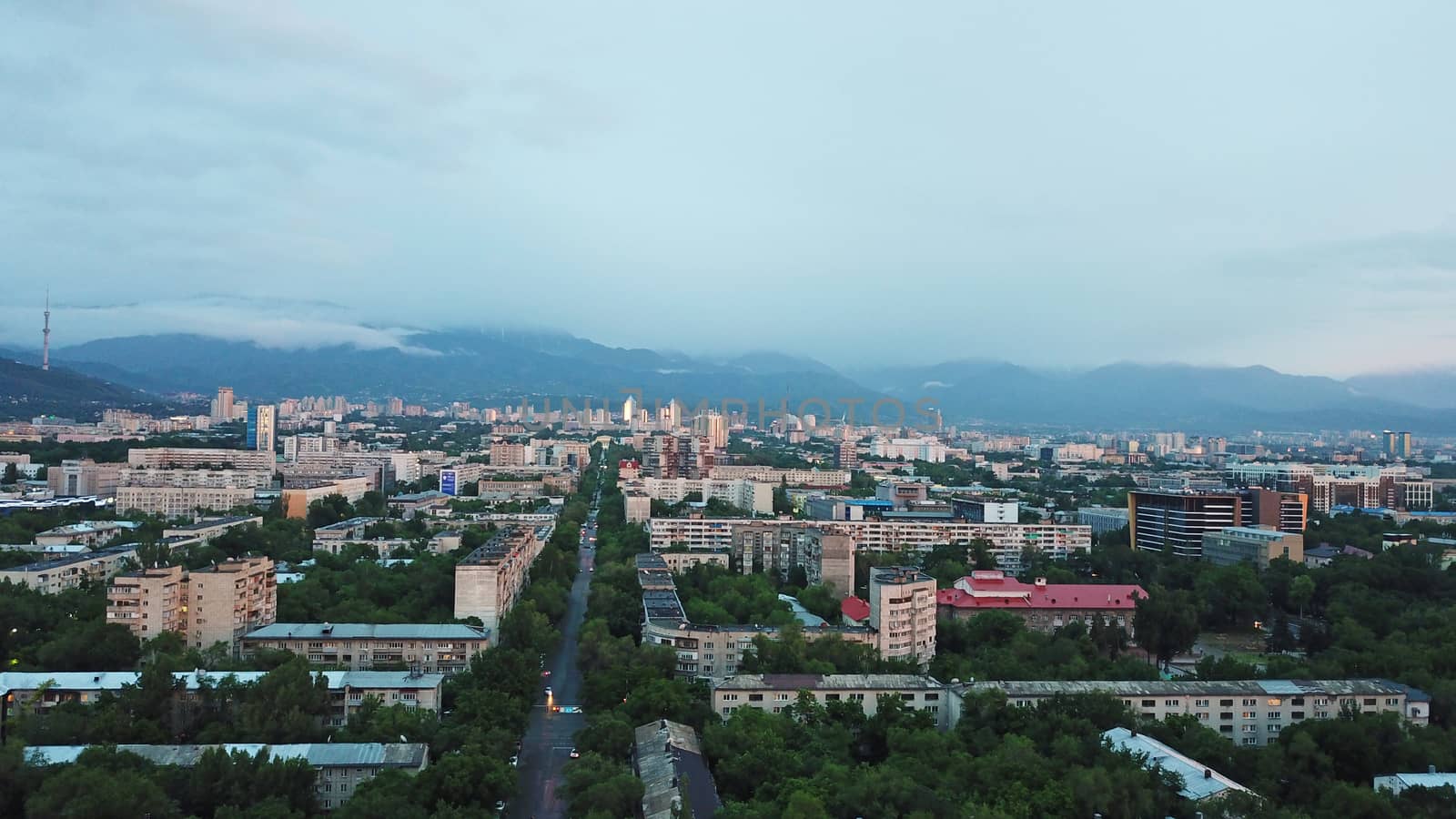 Clouds over the mountains and the city of Almaty at sunset. A lot of green trees, cars driving on the road, high mountains can be seen in the distance. On the hill stands the Kok Tobe TV tower.