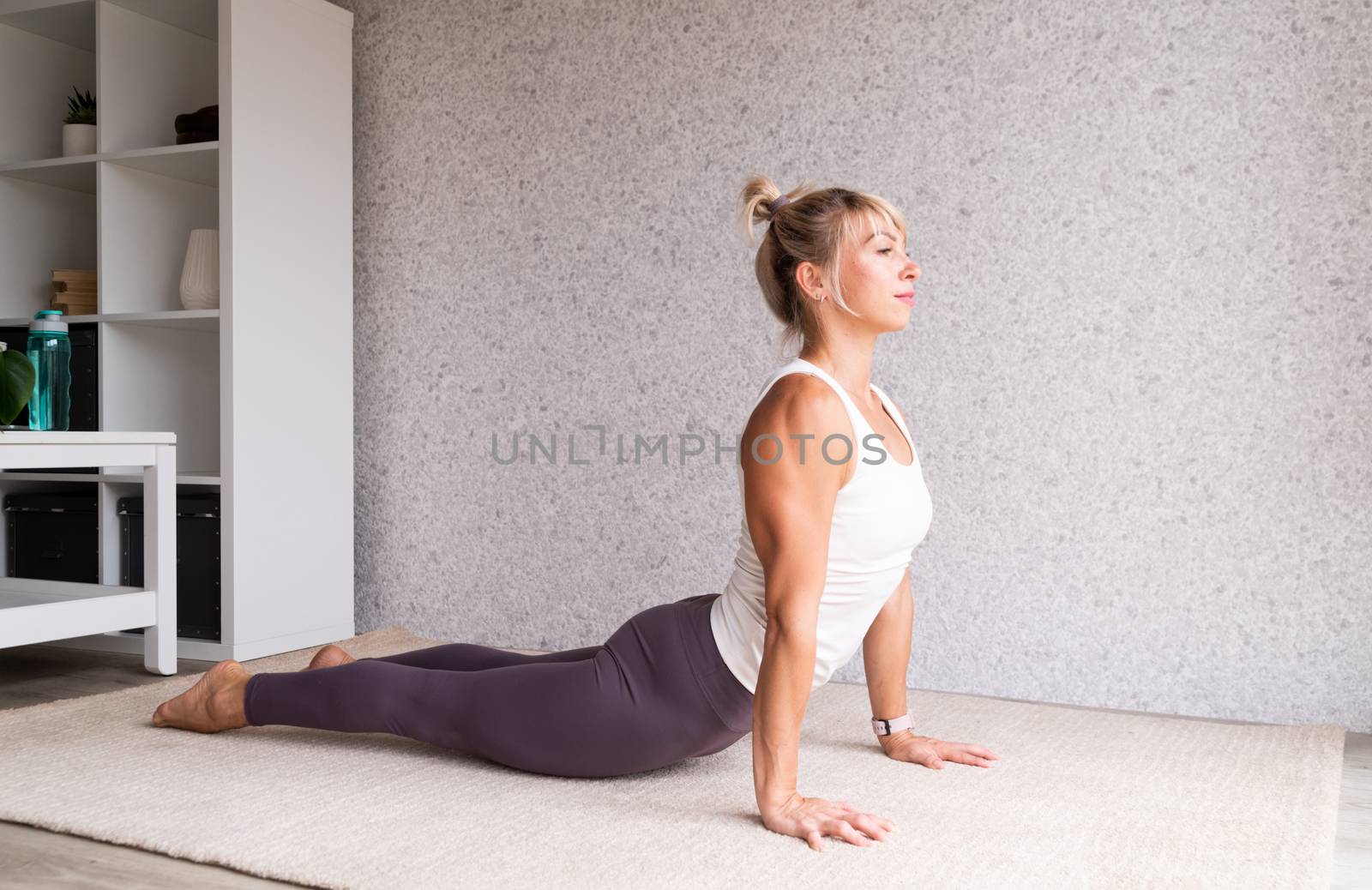 Young attractive woman practicing yoga, wearing sportswear, white shirt and purple pants, indoor full length, gray background by Desperada