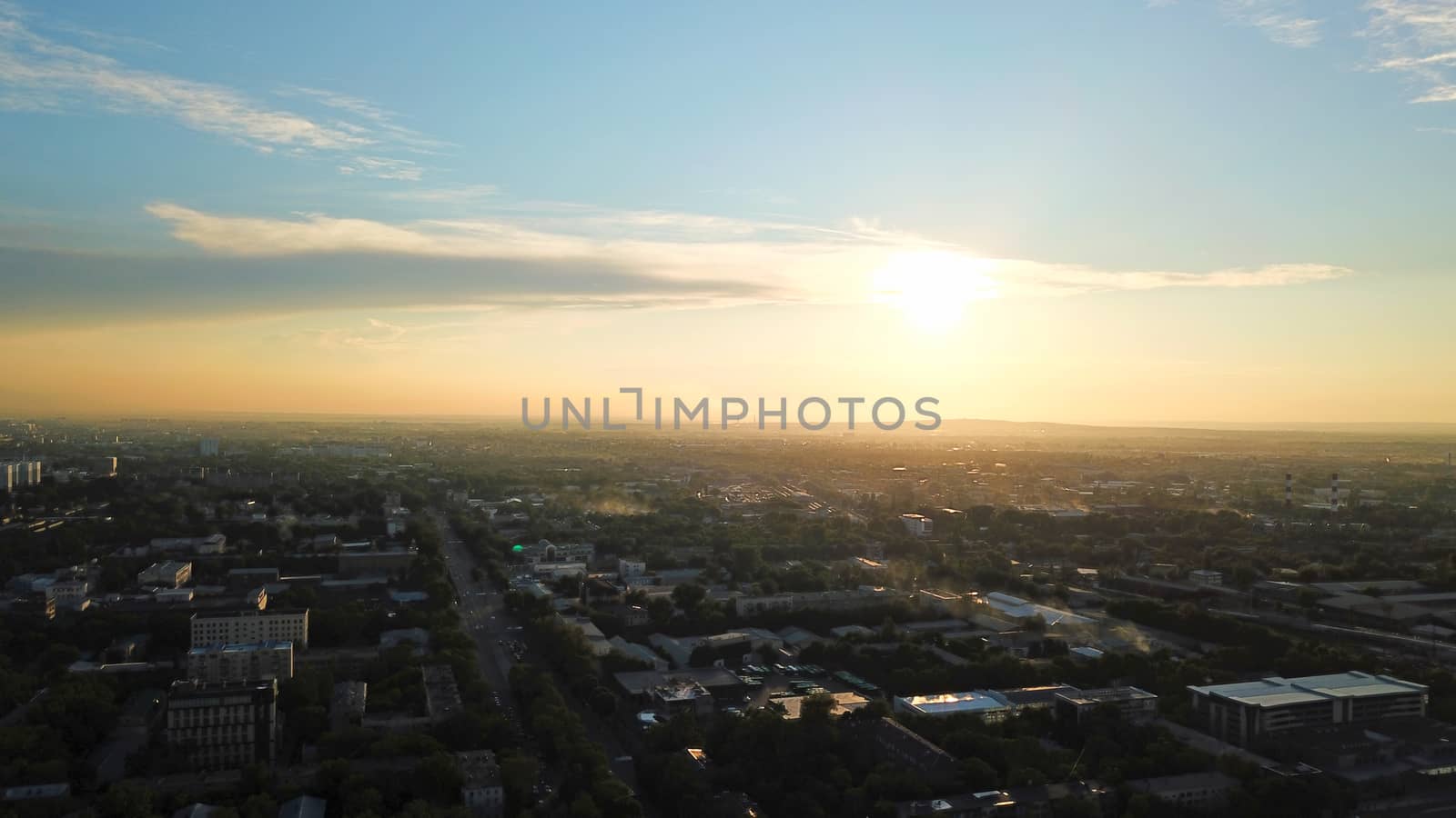 Bright color sunset over the city of Almaty. Huge clouds over the mountains and the city shimmer from bright blue to yellow and dark blue. Tall houses and green trees, cars driving on the roads.