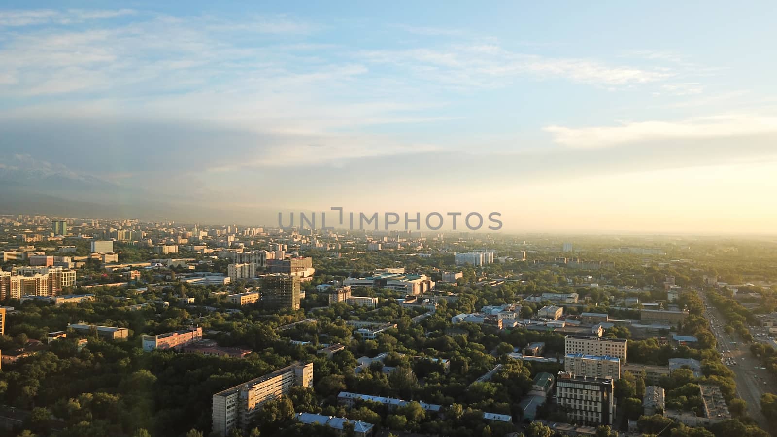 Bright color sunset over the city of Almaty. Huge clouds over the mountains and the city shimmer from bright blue to yellow and dark blue. Tall houses and green trees, cars driving on the roads.