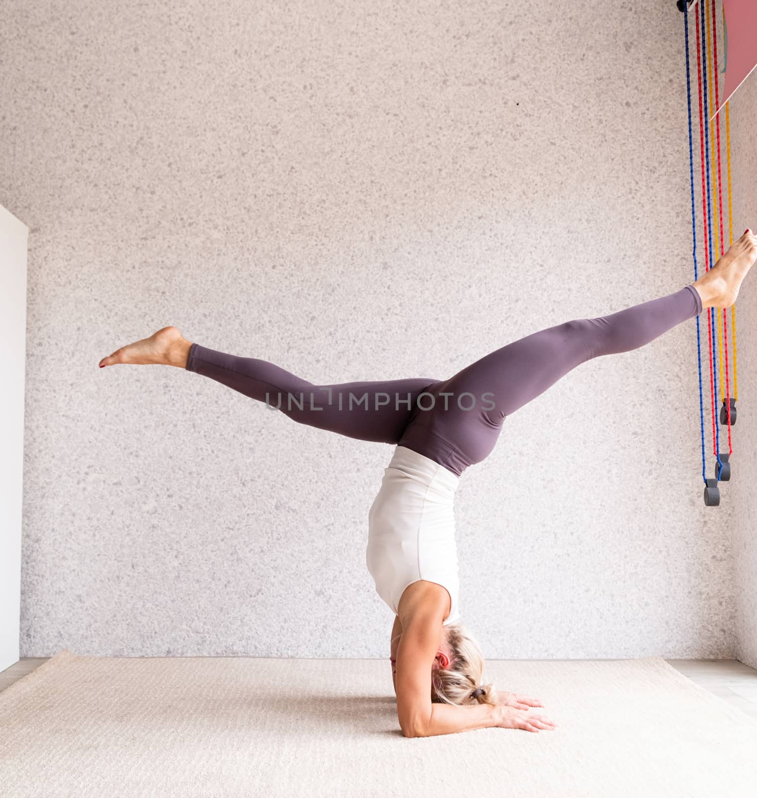 Young attractive woman practicing yoga, wearing sportswear, white shirt and purple pants, indoor full length, gray background by Desperada