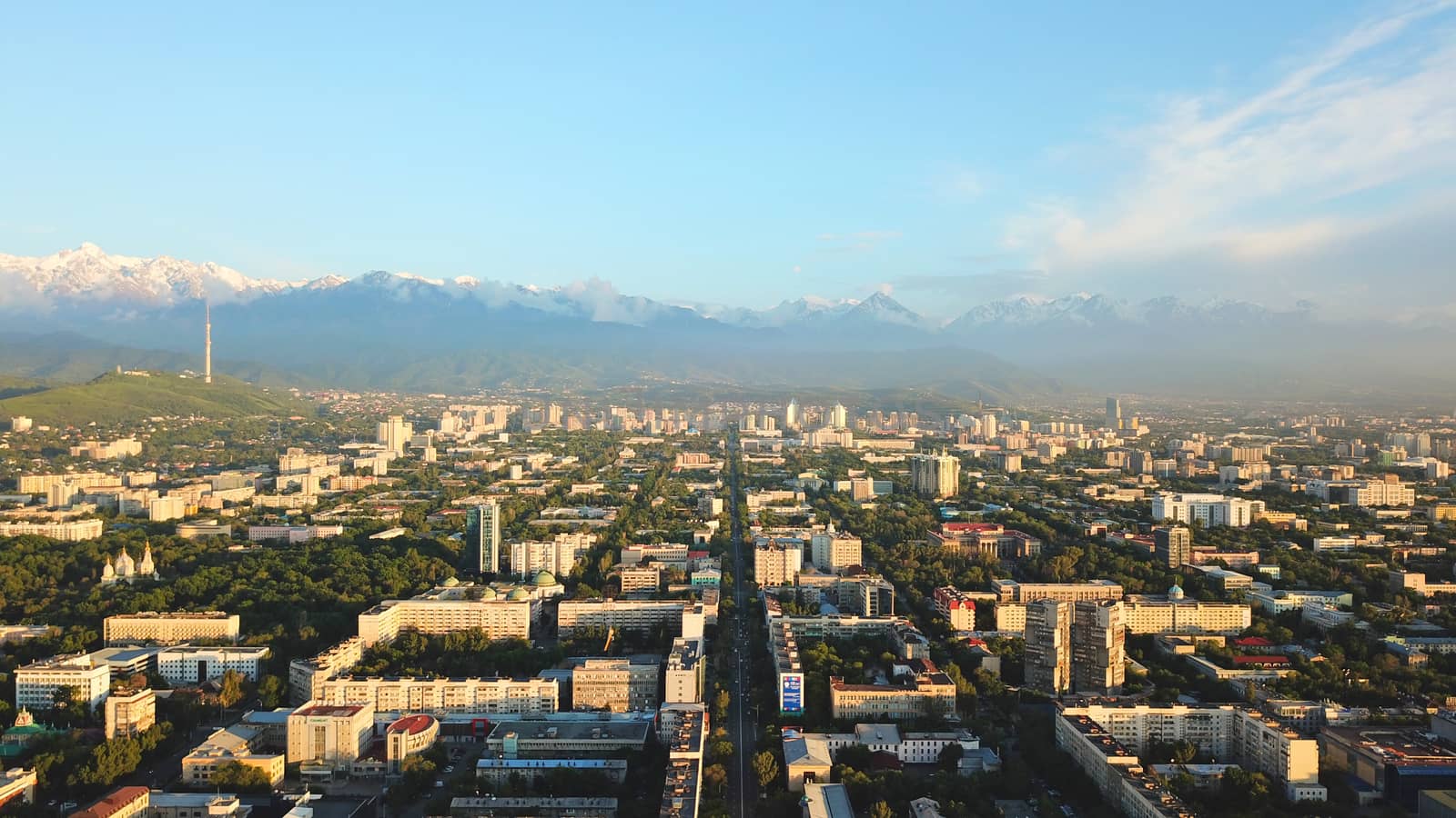 Bright color sunset over the city of Almaty. Huge clouds over the mountains and the city shimmer from bright blue to yellow and dark blue. Tall houses and green trees, cars driving on the roads.