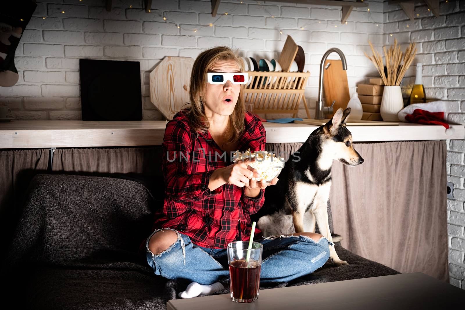 Movie night. Young woman in 3D glasses sitting on the sofa with her dog watching movies at home at night at christmas