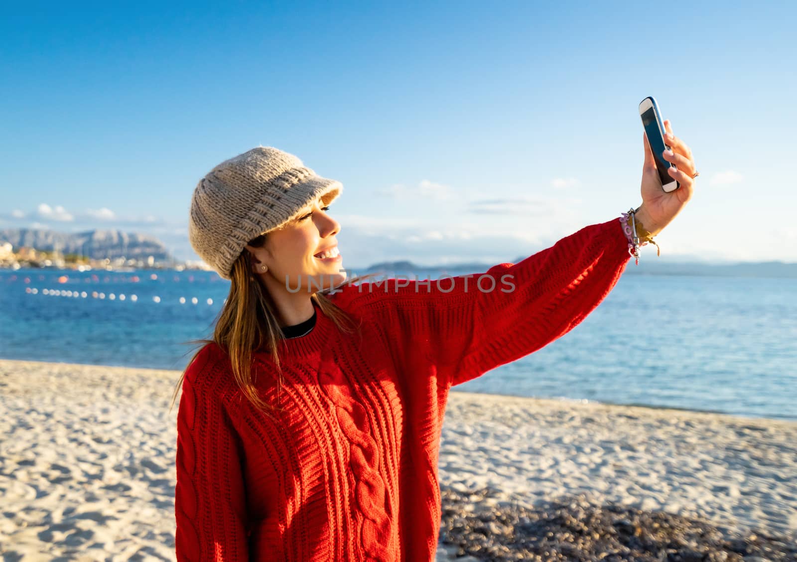 Beautiful young caucasian woman solo traveller make a self portrait herself on the beach at sunset or dawn - Alone female person smiling looking at the phone doing a video call in winter sea vacation by robbyfontanesi