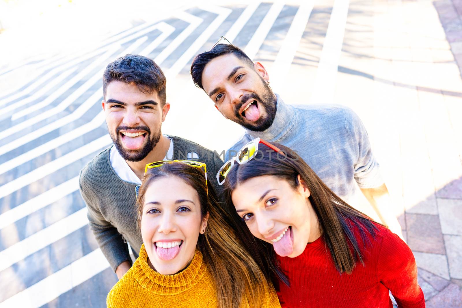 Two Caucasian couple looking at the camera from the bottom smiling and making mouths open faces sticking out their tongues - Friends having fun outdoor doing selfie with camera on top - Focus on men